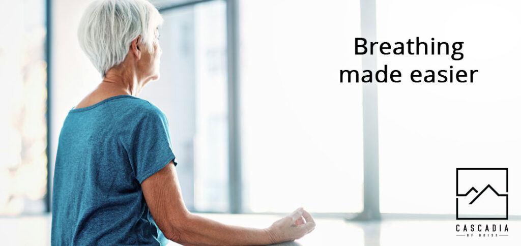 The back and side profile of a senior women standing and looking out a window while resting her arms on a deep window sill. The text "Breathing made easier" and the Cascadia of Boise logo are on the picture.