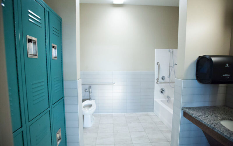 A bathroom used for Occupational Therapy. A tile floored bathroom with two rows of lockers on the left and a sink with a counter across from them. A toilet and tub with grab bars are at the far end of the room.