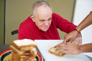 A senior man sitting in a wheelchair with a pleasant look on his face is using his left hand to apply condiments to a piece of bread. The hands of a therapist are holding the slice of bread.