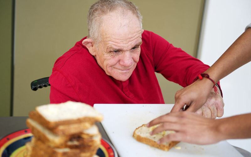 A senior man sitting in a wheelchair with a pleasant look on his face is using his left hand to apply condiments to a piece of bread. The hands of a therapist are holding the slice of bread.