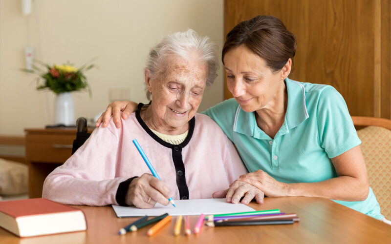 A smiling senior woman is sitting at a desk with a colored pencil in her right hand and a piece of paper in front of her. To her left is a smiling young woman with her right arm around the woman's shoulder and her left hand placed gently on the woman's left hand.