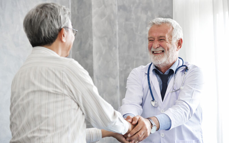 A senior doctor is smiling wide as he shakes the hand of a senior man.