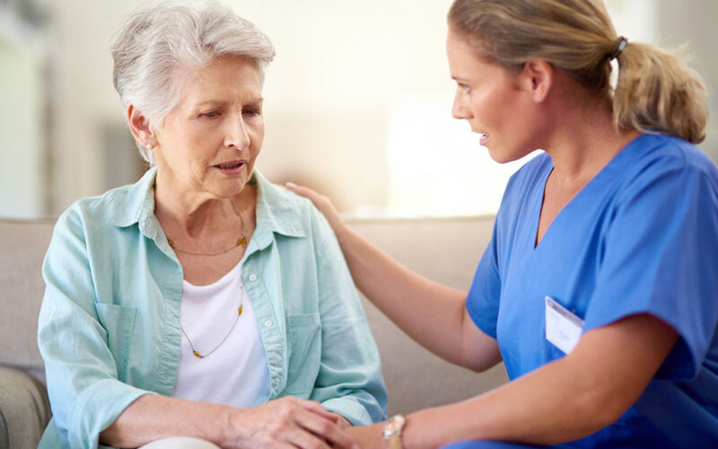 A senior woman is sitting in a chair talking. To her left a female therapist is also talking and has her right arm placed gently on the woman's left shoulder and her left hand gently grasping the older woman's right hand as they work on speech therapy skills.