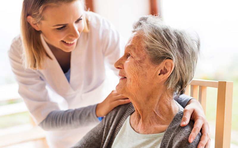 A smiling senior woman is sitting in a chair looking up at a younger woman who is smiling down at her. The younger woman has her left arm placed gently on the older woman's shoulder and her right hand on her right shoulder.