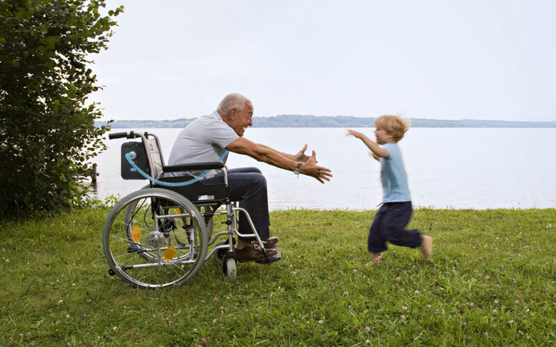 A smiling senior man sitting in a wheelchair with his arms outstretched in front of him to greet a young boy who is enthusiastically running toward the man. The pair are in a grassy field on the edge of a calm lake on a slightly hazy day.