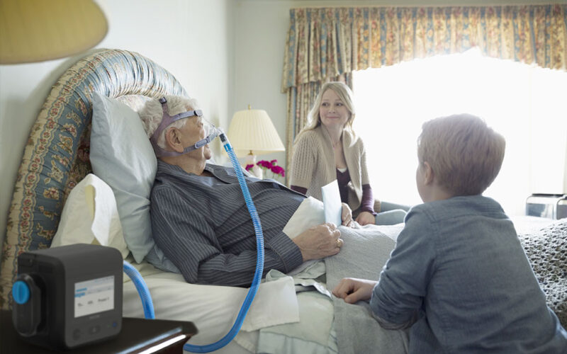 A senior man wearing a respiratory ventilator placed over his nose while lying in a bed in a well-decorated room. A smiling young woman and a younger boy are beside the man, leaning on his bed.